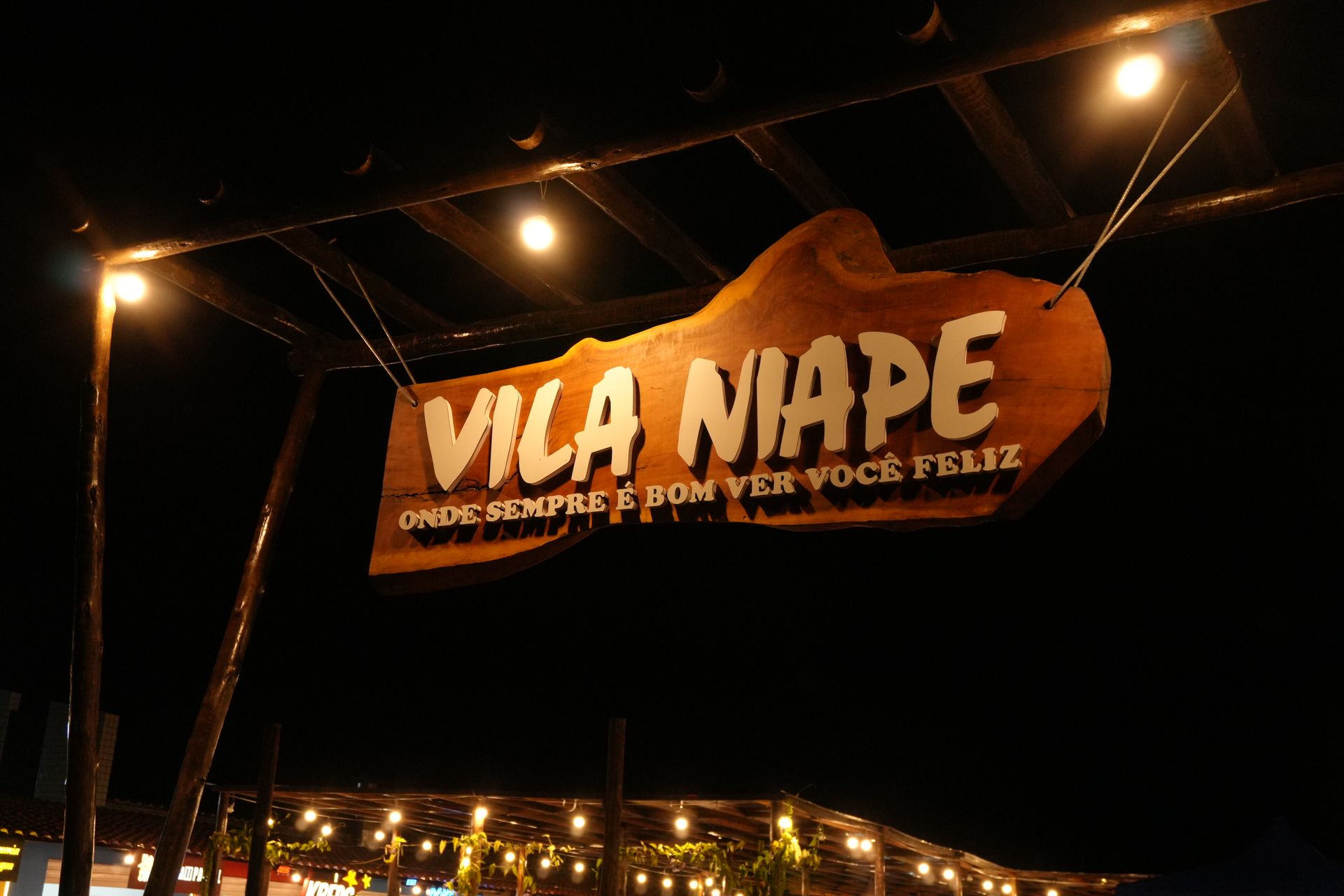 Placa de madeira à noite com a inscrição "Vila Niape" em branco e luzes de cordão abaixo.