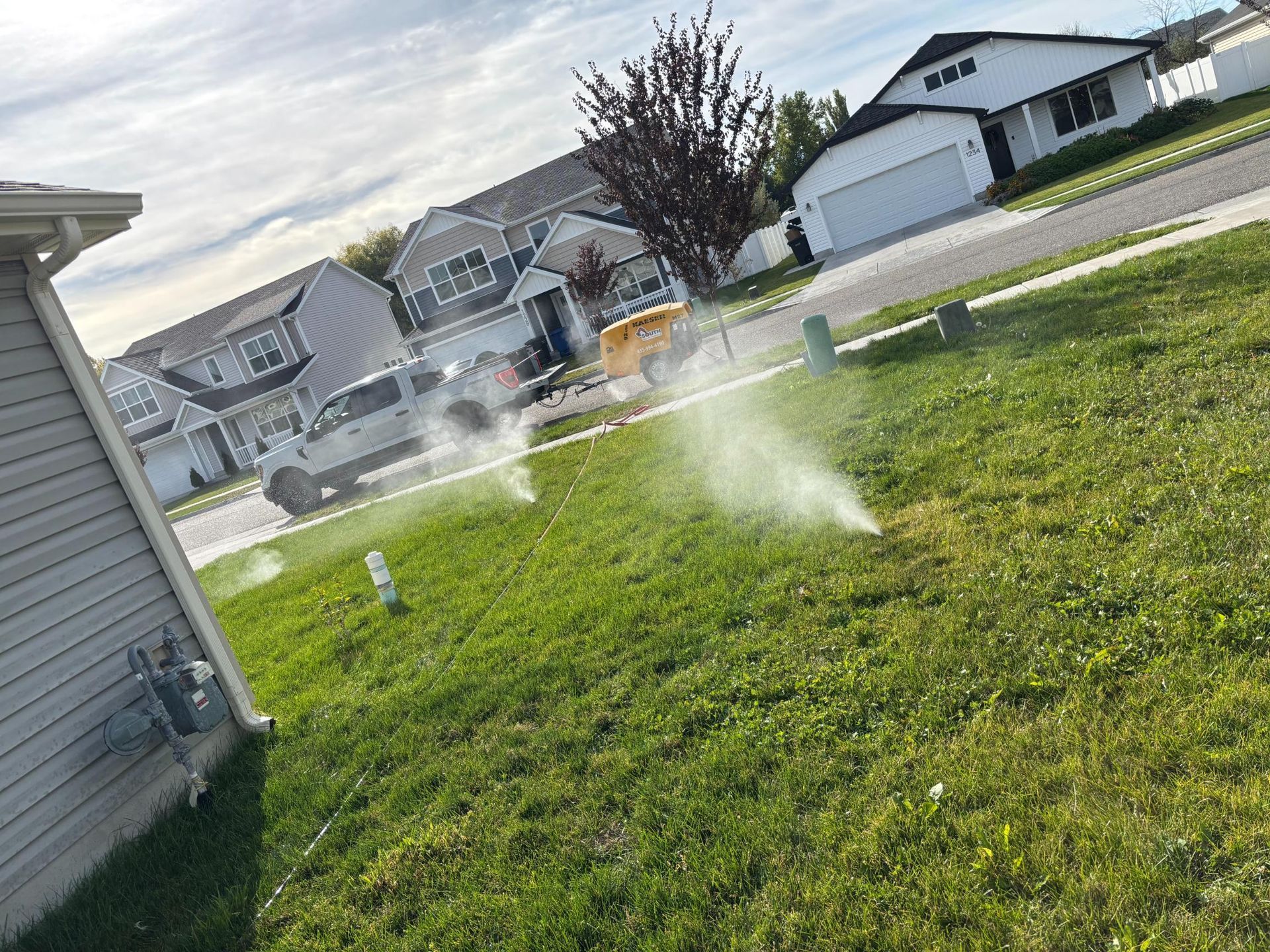 Sprinklers watering a green lawn in a suburban neighborhood, with houses and a van in the background.