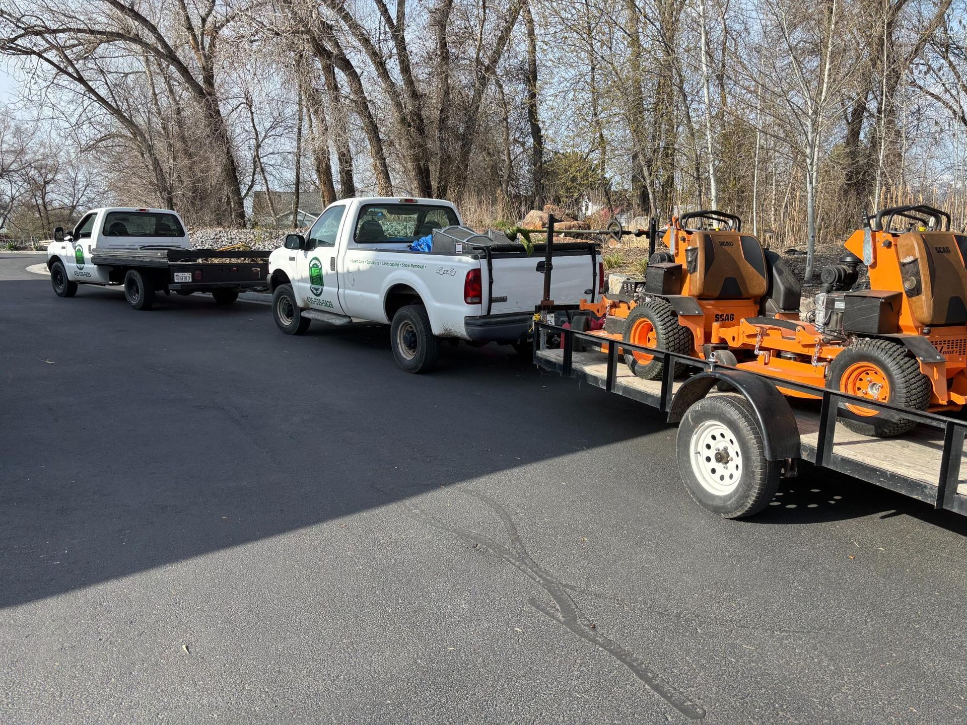 Two white trucks towing a trailer with two orange lawn mowers on a paved road.
