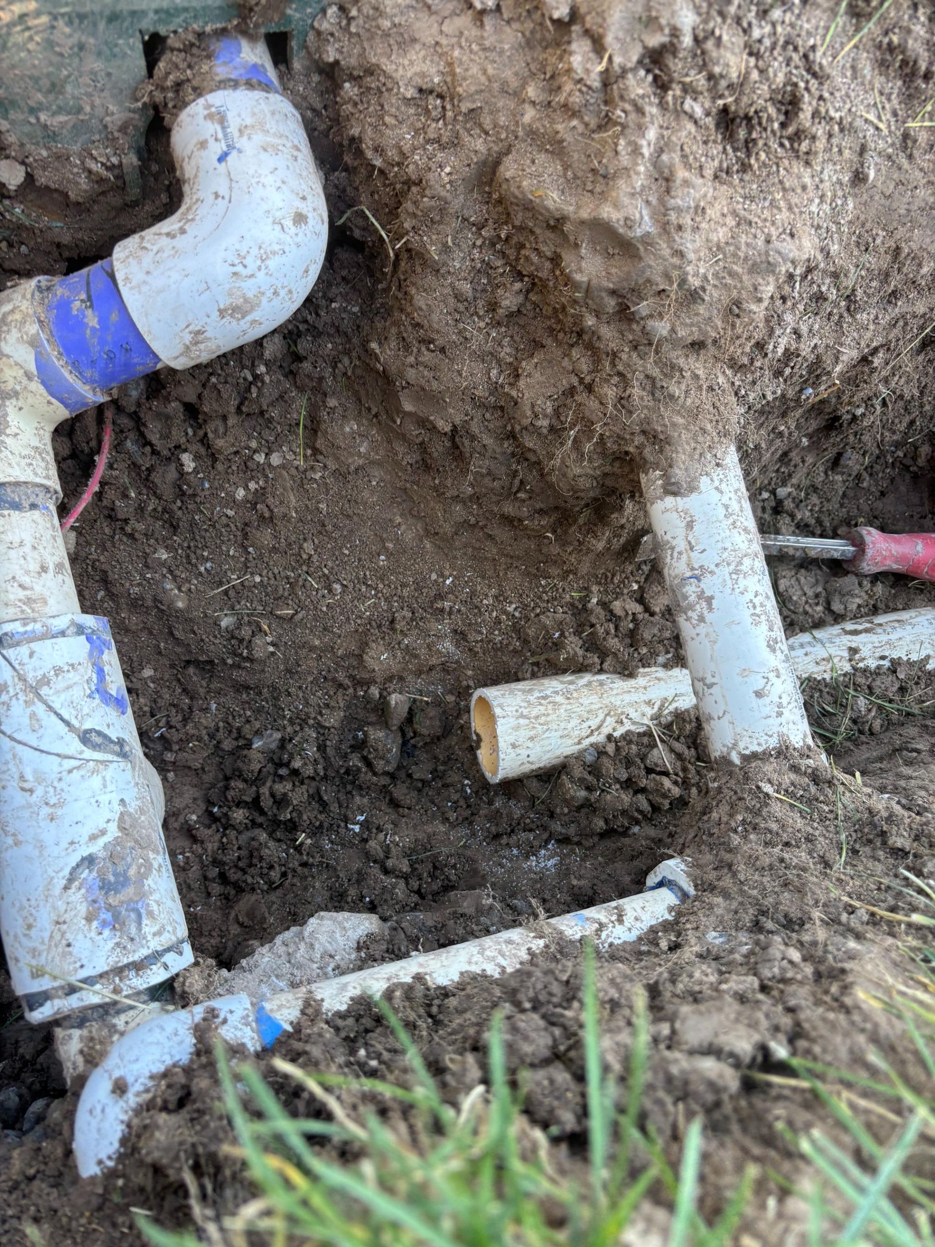 Irrigation system plumbing in a dirt pit. White PVC pipes with blue fittings, dirt and a trowel are visible.