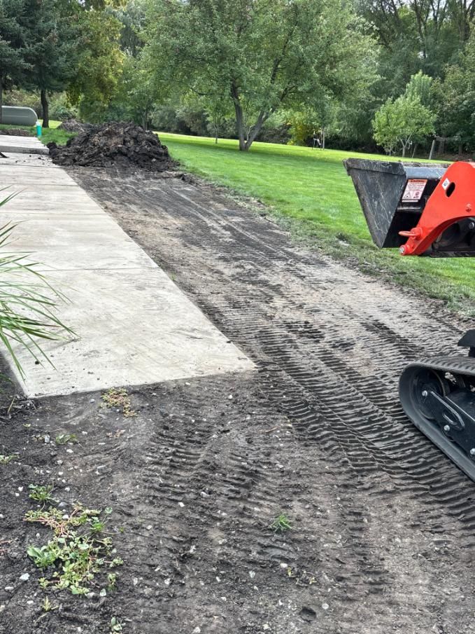 A concrete walkway next to a dirt path and grass. A small earth-moving machine sits at the right.