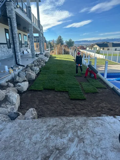 Two people laying sod near a pool, a house, and a rock border. Blue sky and mountains.
