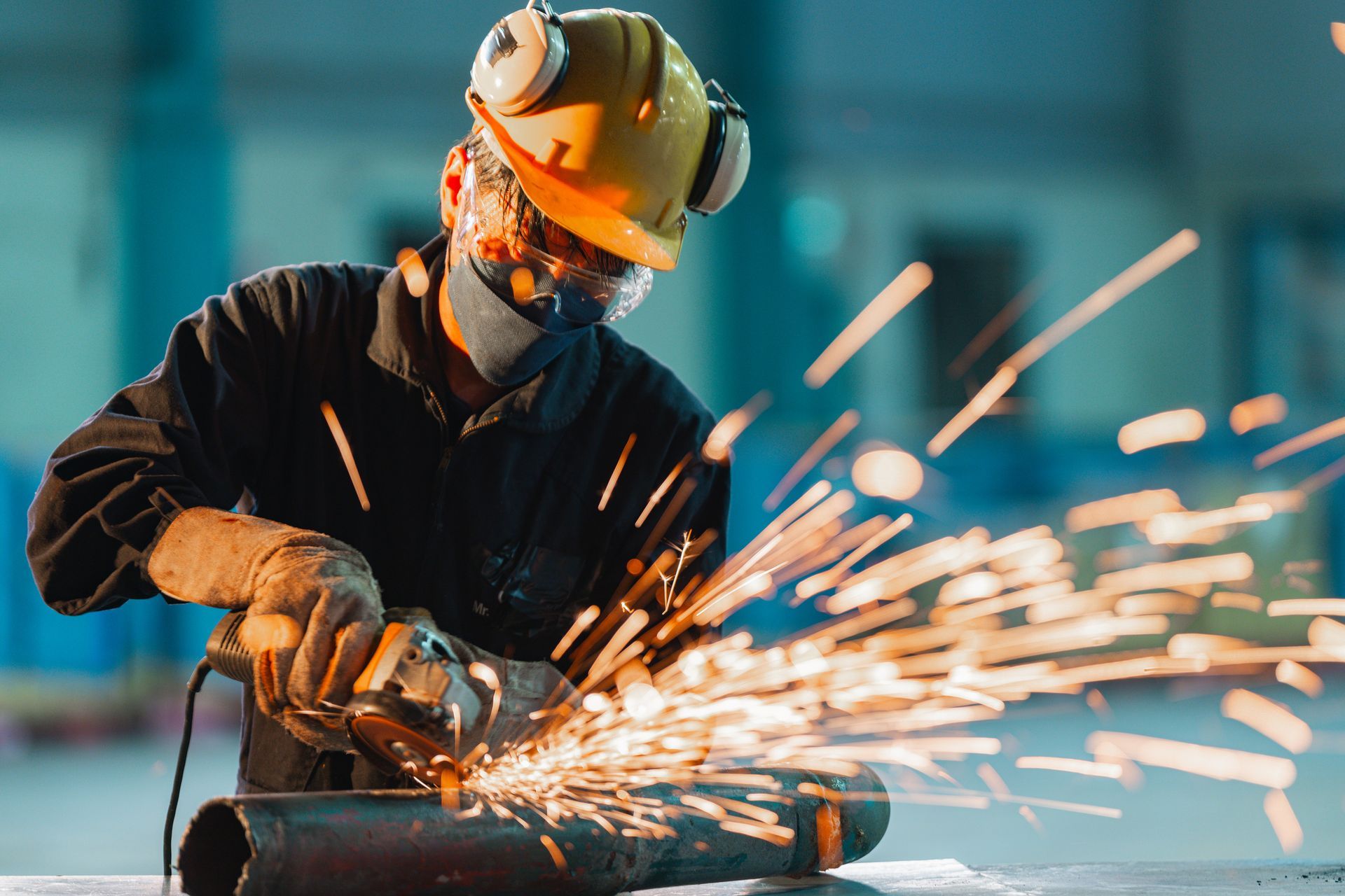 Worker in a hard hat and mask grinds metal, creating sparks in a workshop.