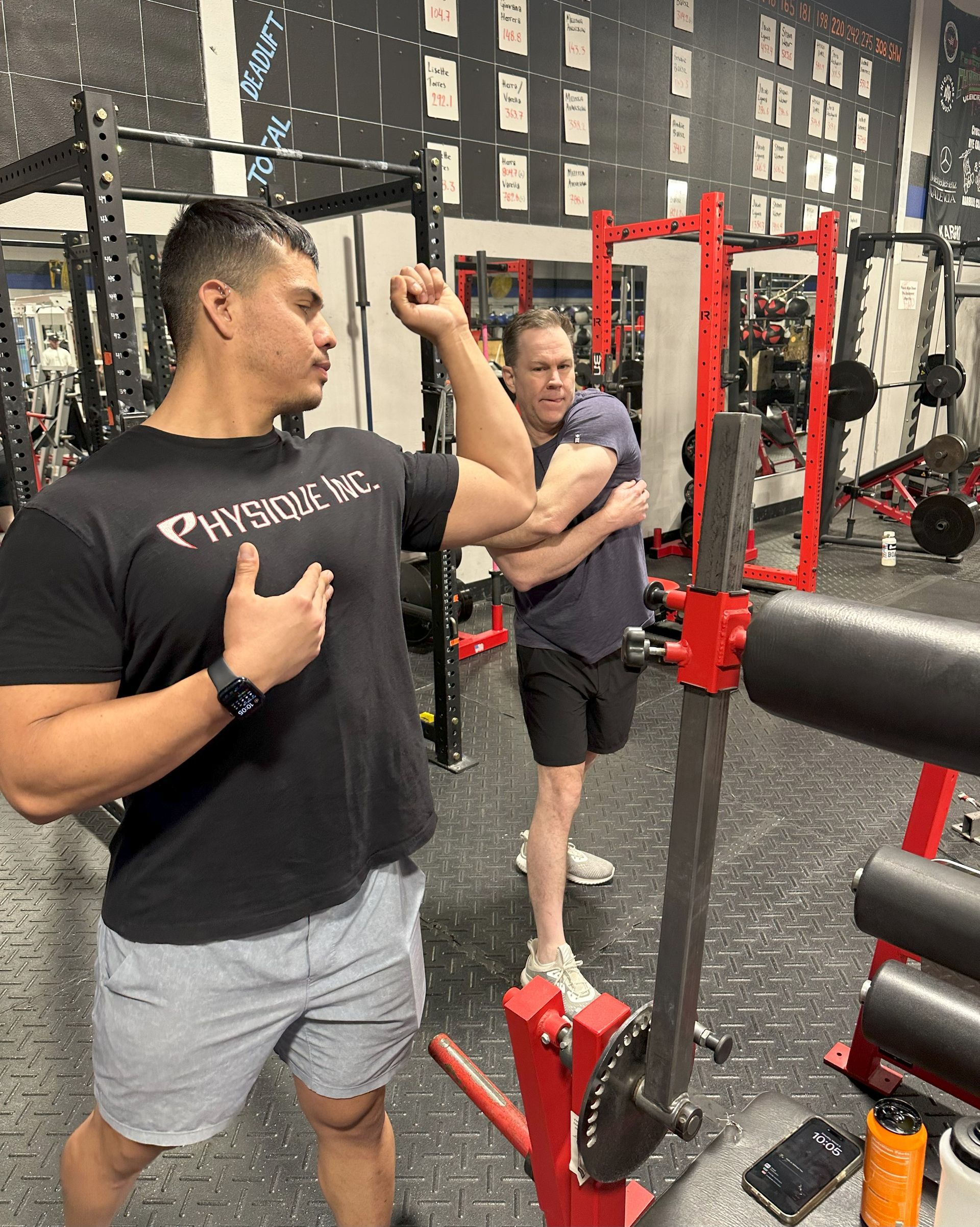 A man is flexing his muscles in a gym while another man looks on.