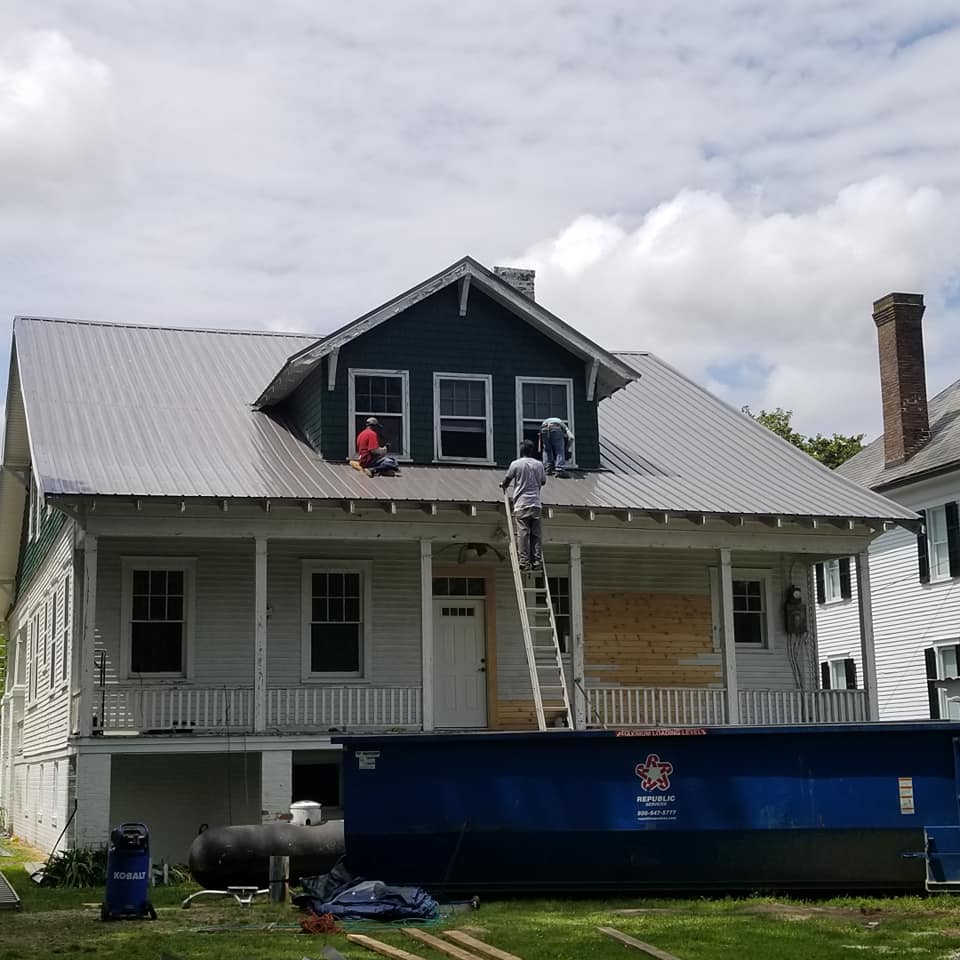 Two people on a house roof, one on a ladder. Metal roof, white siding, blue dumpster. Cloudy sky.
