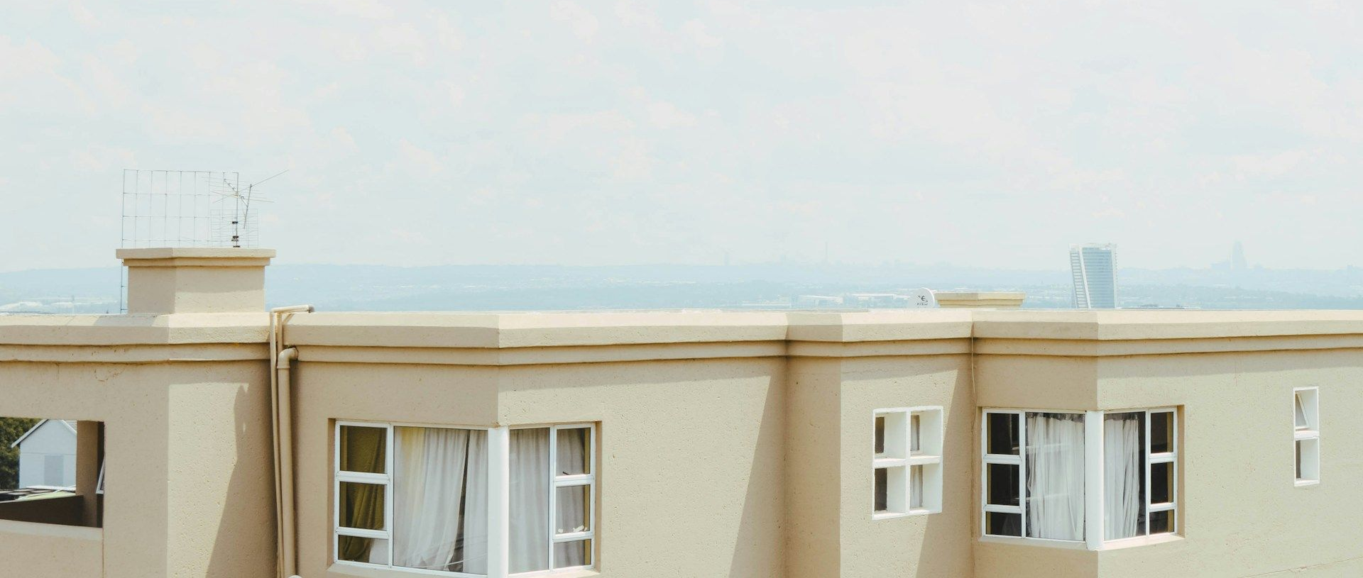 A beige building with white-framed windows against a hazy sky. Distant city skyline and antenna on the roof.