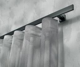 Tende bianche trasparenti appese a un bastone grigio scuro dai bordi squadrati, montato su una parete grigio chiaro.