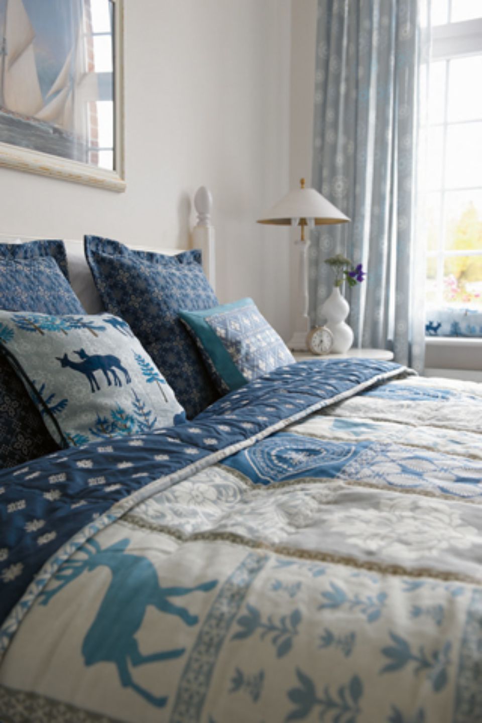 Letto con lenzuola e cuscini a fantasia blu, testiera bianca e finestra con tende.