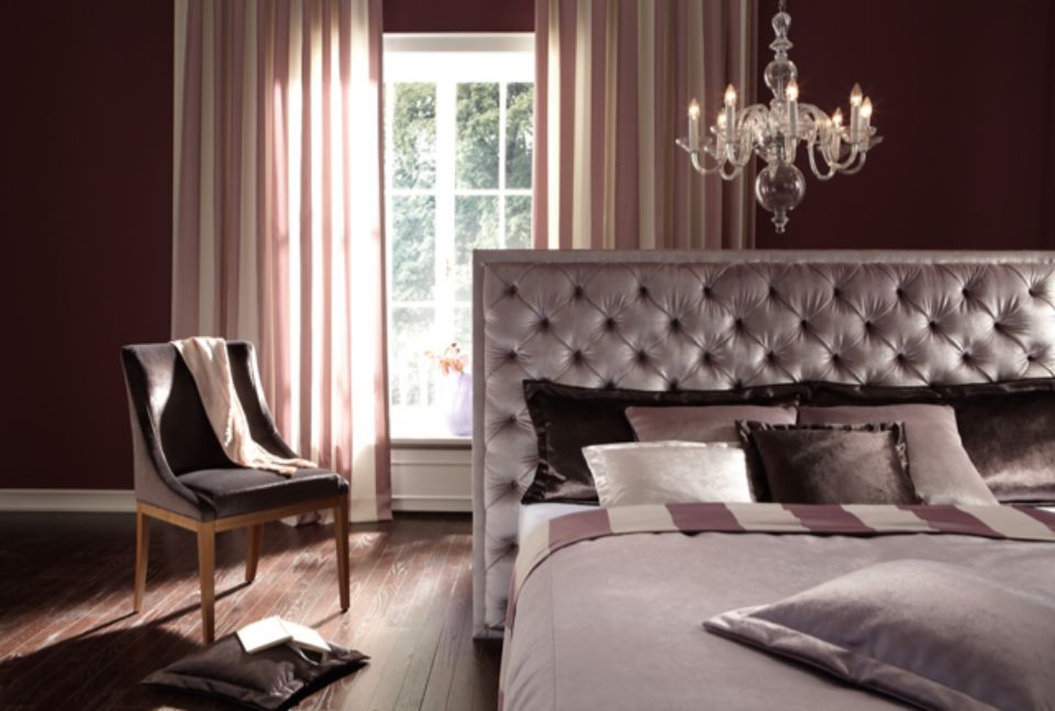 Camera da letto con ampia testiera imbottita, pareti color bordeaux, tende a righe, lampadario e una sedia con coperta.