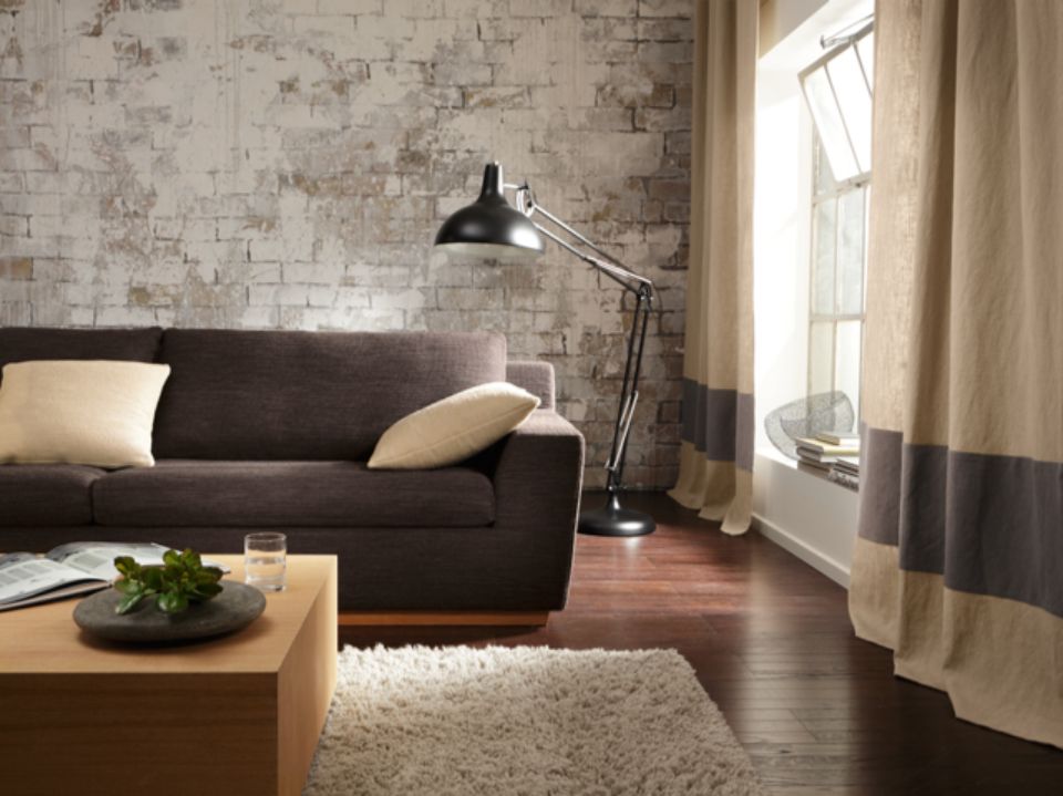 Soggiorno con divano marrone, parete in mattoni bianchi anticati, lampada da terra e tende a righe.