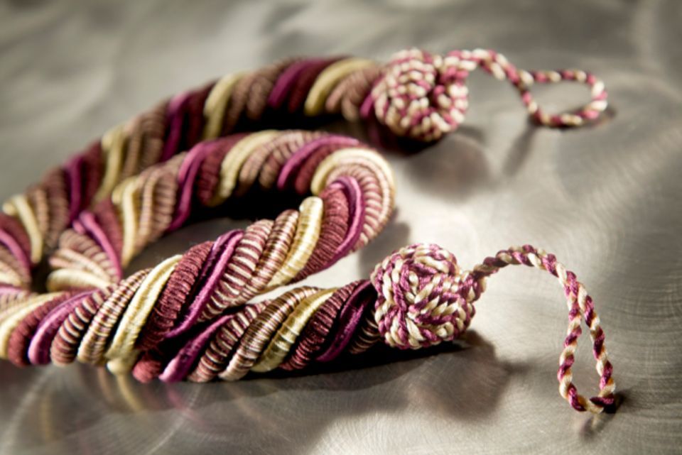 Corda intrecciata nei toni del viola, del beige e del bianco, con anelli a ciascuna estremità, appoggiata su una superficie.