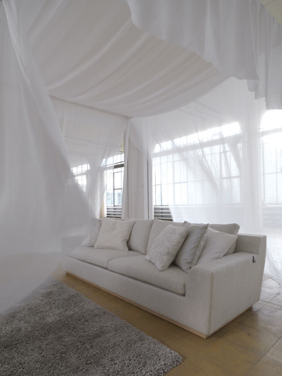 Divano bianco in una stanza con tessuto bianco drappeggiato e grandi finestre; atmosfera morbida e ariosa.