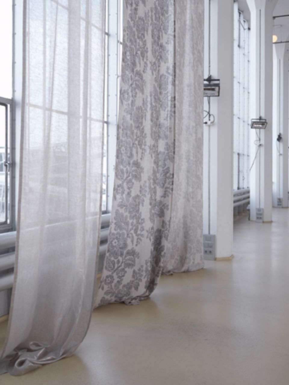 Lunghe tende trasparenti davanti a una grande finestra. Pavimento beige chiaro. Colonne bianche sullo sfondo.