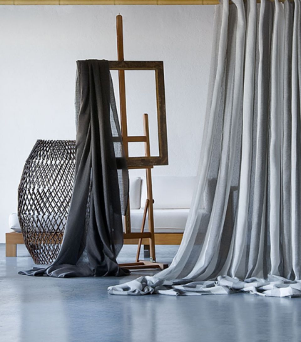 Una tenda grigia drappeggiata e un tessuto su un cavalletto; un tavolino di vimini; un divano bianco.