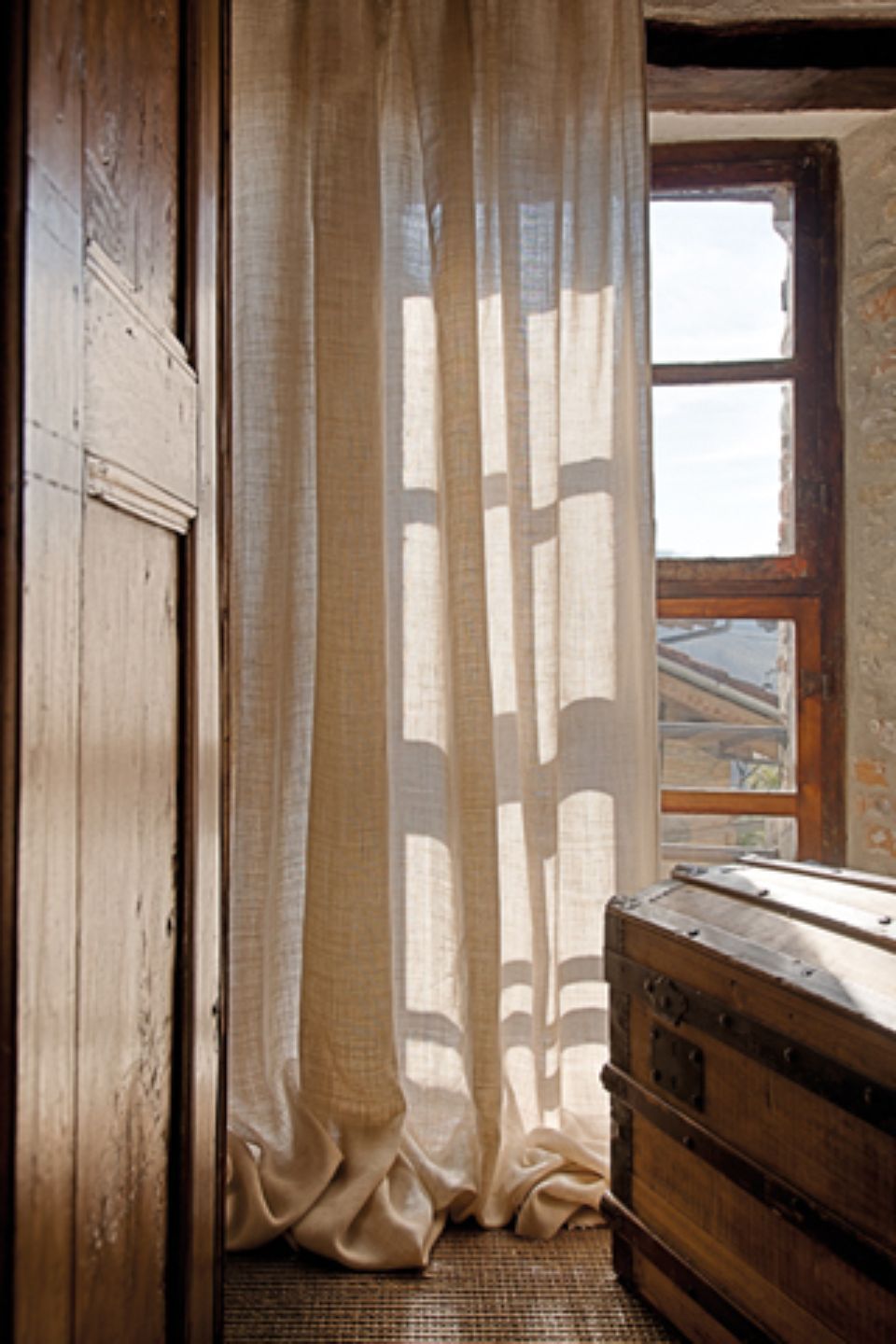 Tende di lino drappeggiate davanti a una finestra, una cassapanca di legno e una porta.