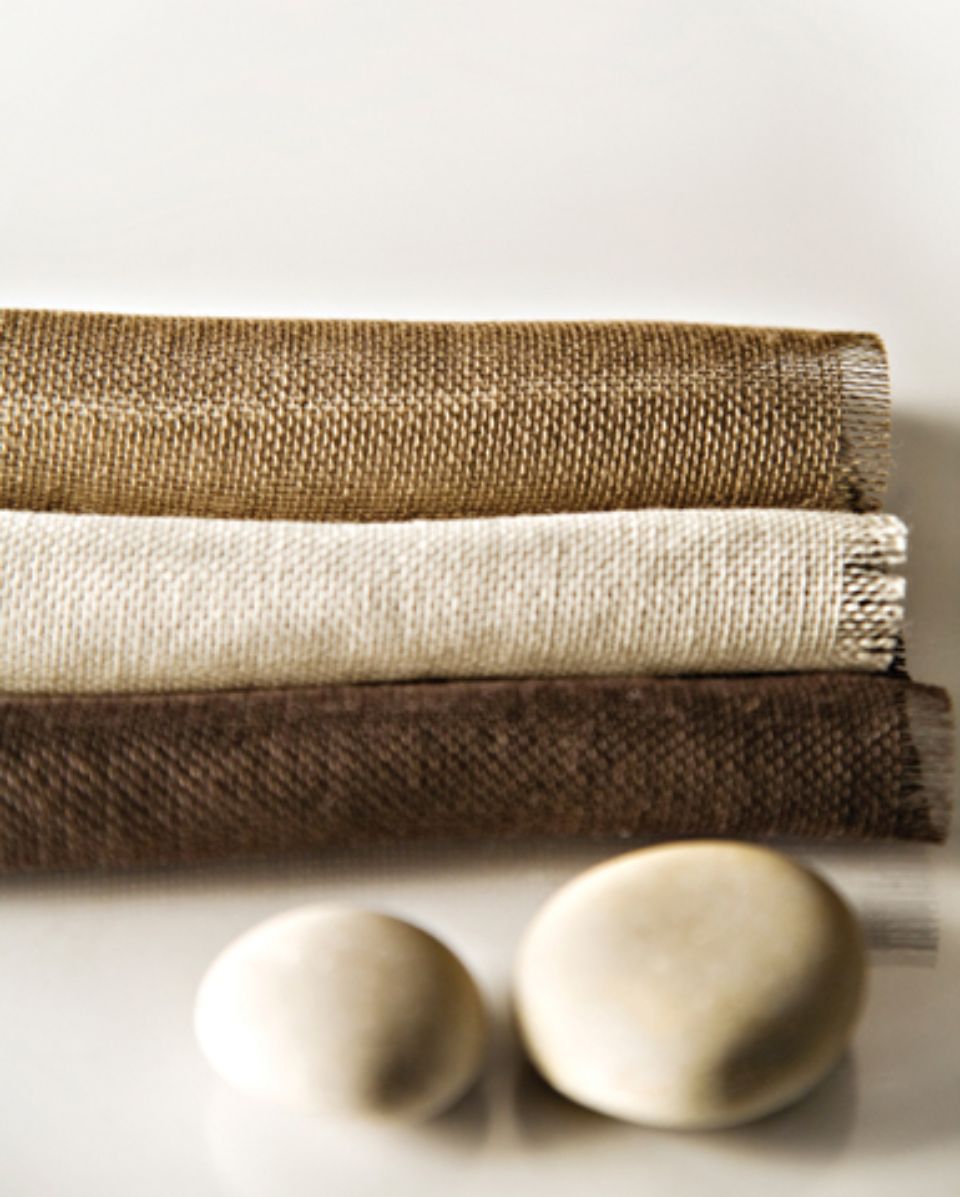 Tre campioni di tessuto piegati nei colori beige, crema e marrone con due pietre lisce su una superficie bianca.