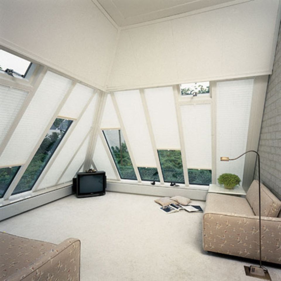 Stanza angolare con pareti bianche, ampie finestre, TV, divano e moquette. La luce del sole entra a fiotti.