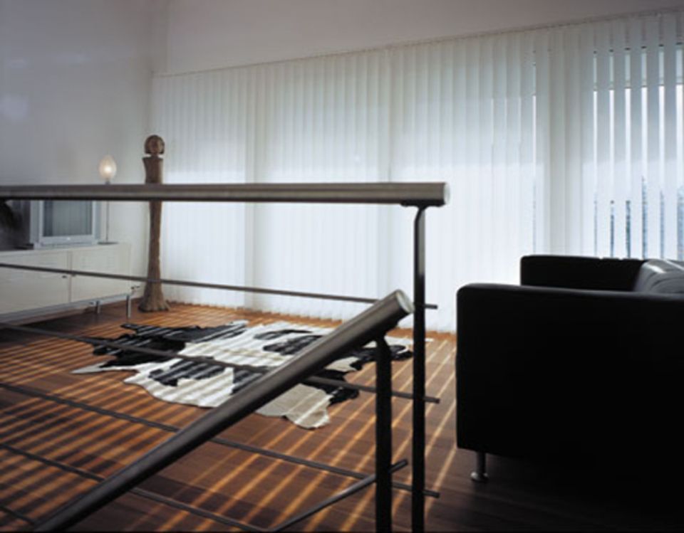 Interni moderni con ringhiera, tappeto in pelle di mucca e tende verticali che proiettano ombre sul pavimento in legno.