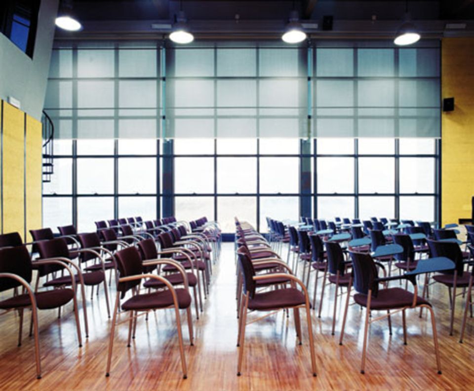 Una sala conferenze vuota con file di sedie di fronte a una grande finestra. Pavimento in legno e parete gialla.