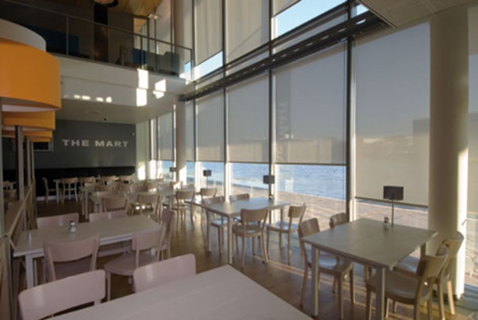 Interno di un ristorante luminoso, con tavoli, sedie e grandi finestre con tende chiuse. È visibile l'insegna