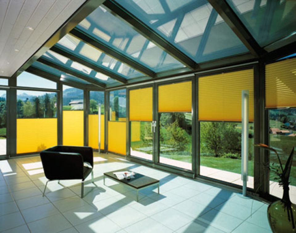 Veranda con tende gialle, tetto in vetro, pavimento piastrellato, sedia nera e vista sugli alberi e sulla collina.