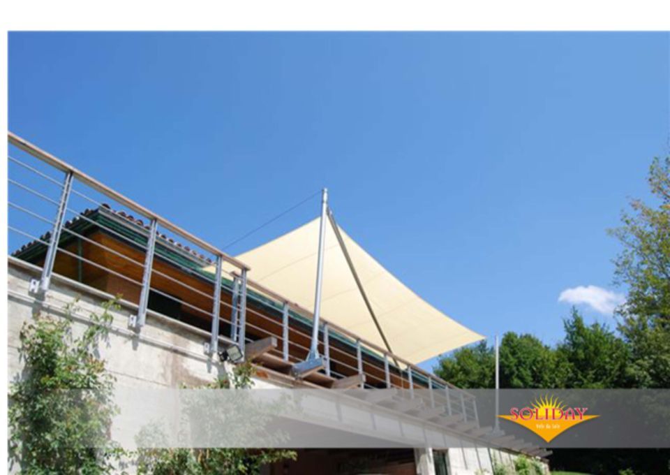 Vela ombreggiante beige sopra un'area salotto all'aperto, sostenuta da un palo, contro un cielo azzurro.