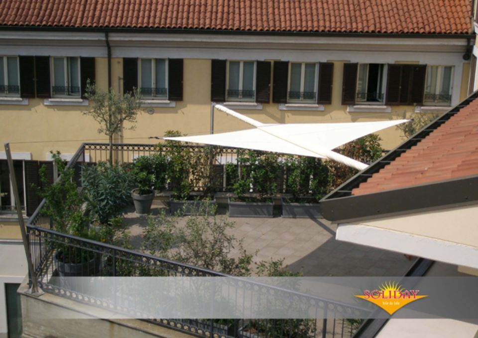 Giardino pensile con vela bianca, ringhiera nera e edificio con pareti gialle e persiane marroni.