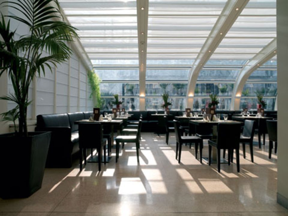 Interno del ristorante con tavoli, sedie e un grande soffitto curvo in vetro. Palma sulla sinistra.