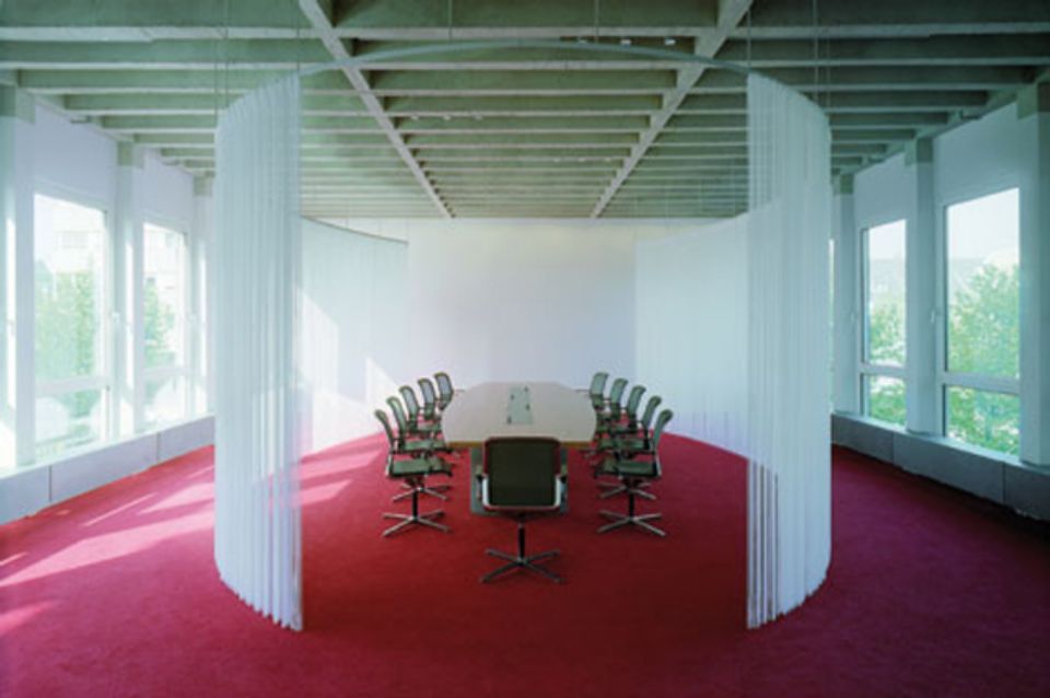 Una sala conferenze con un lungo tavolo, sedie e tappeto rosso, delimitata da divisori verticali bianchi.