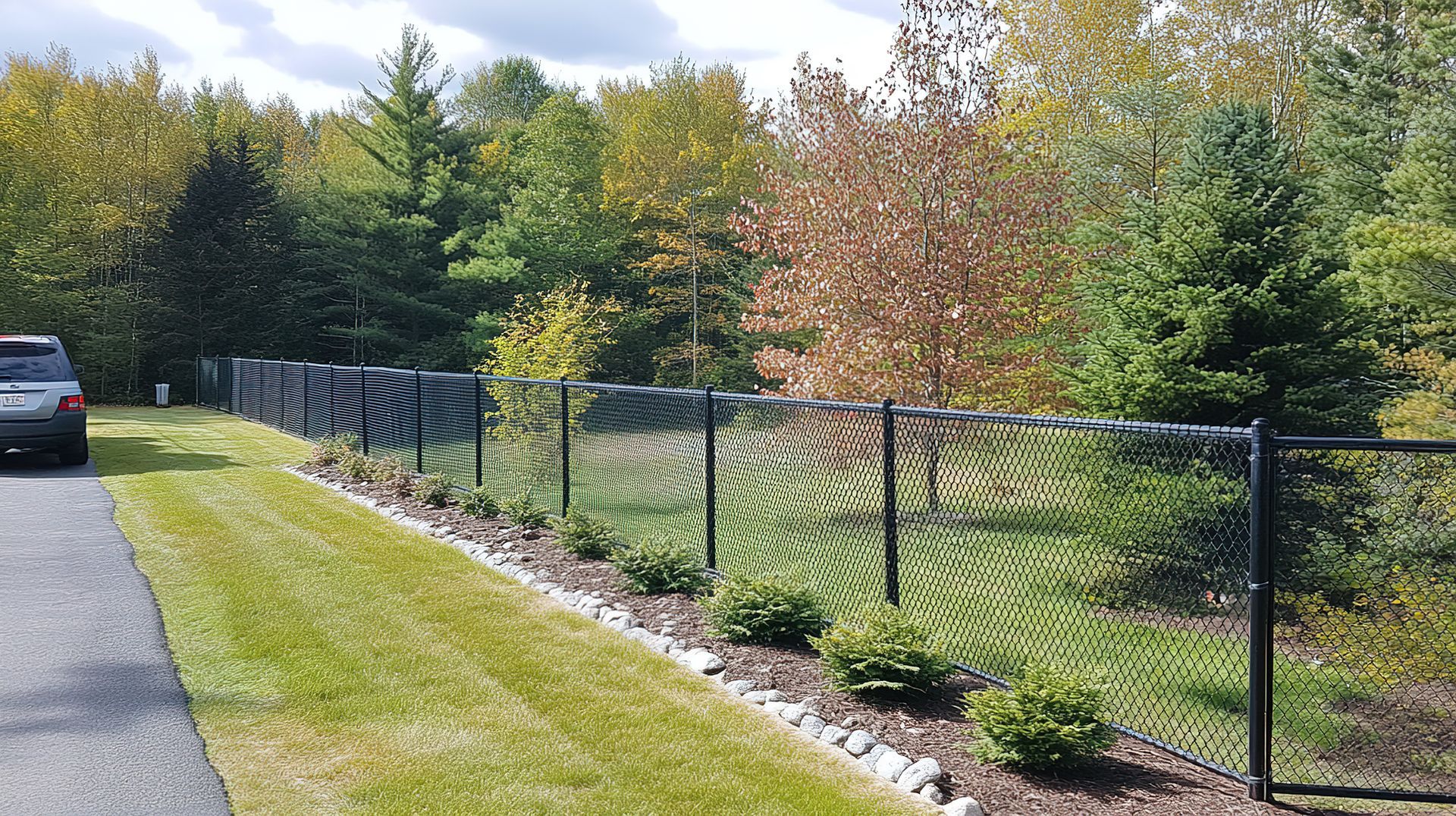 Black chain-link fence along a grassy path beside a road, with trees and shrubs in the background