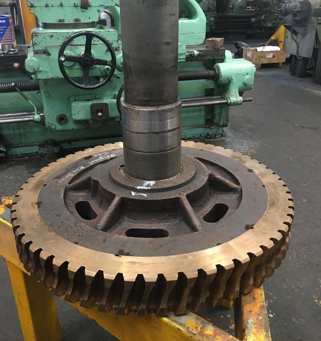 A large, circular, dark-colored gear with slanted teeth mounted on a steel shaft, sitting in a machine workshop.