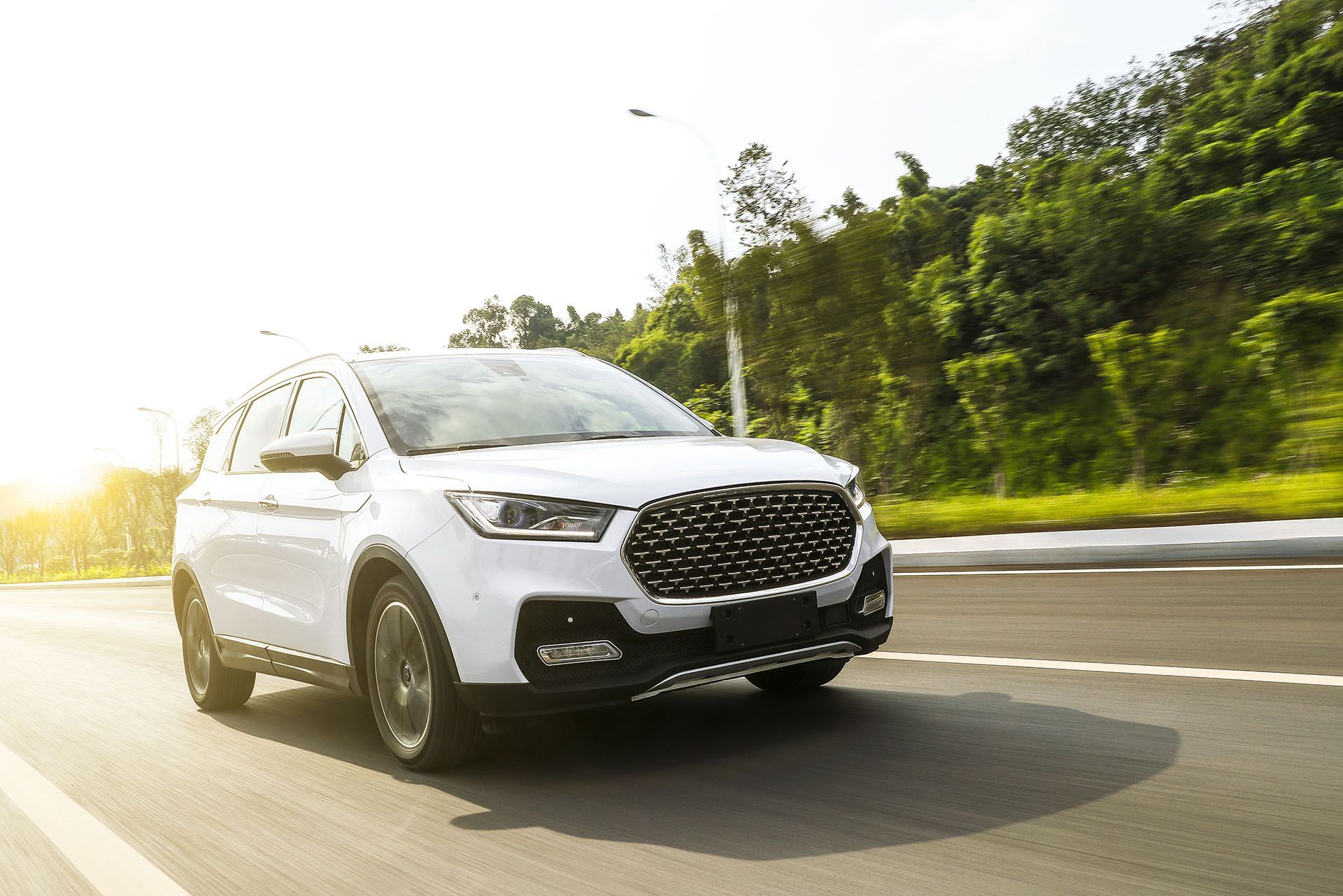 Un SUV crossover bianco percorre una strada asfaltata in una giornata di sole, con una verde collina sullo sfondo.