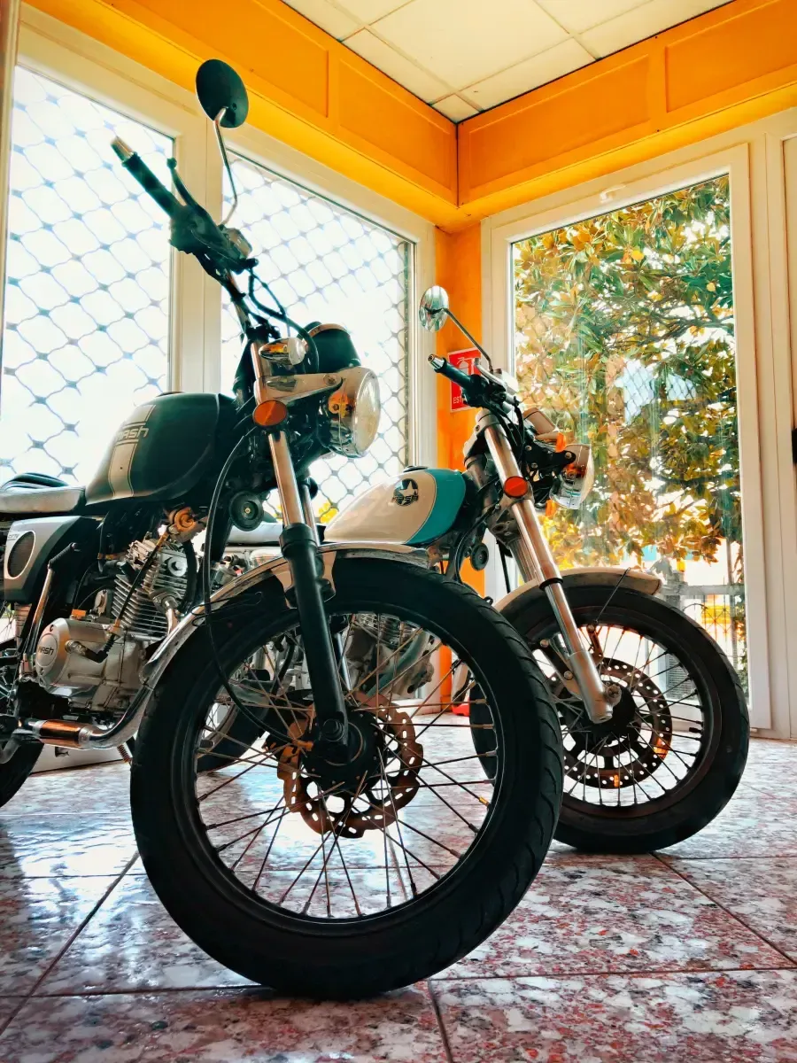 Due motociclette parcheggiate all'interno, su un pavimento piastrellato, vicino a una grande finestra, illuminate da una calda luce naturale.