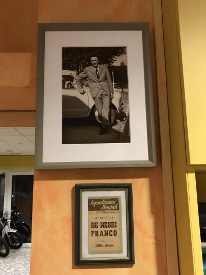 Fotografia in bianco e nero incorniciata di un uomo appoggiato a un'auto, montata sopra un piccolo ritaglio di giornale d'epoca anch'esso incorniciato.