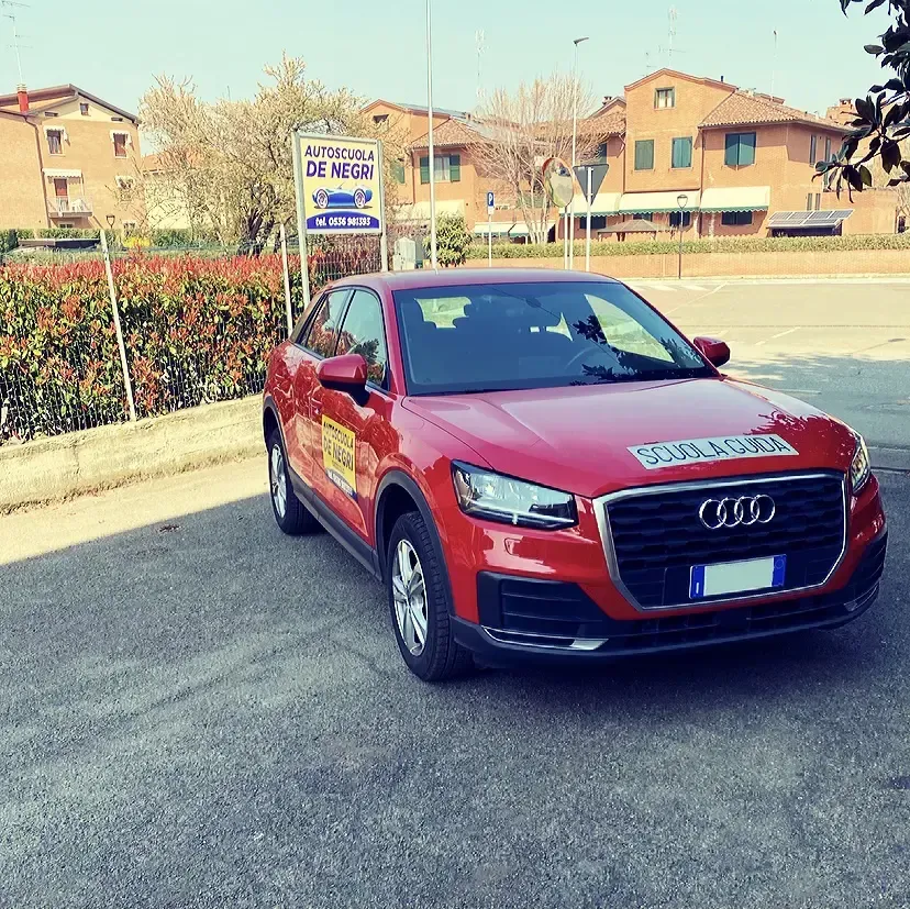 Un'Audi rossa, auto di una scuola guida, parcheggiata su un piazzale asfaltato di fronte a un edificio in mattoni rossi e a un cartello.