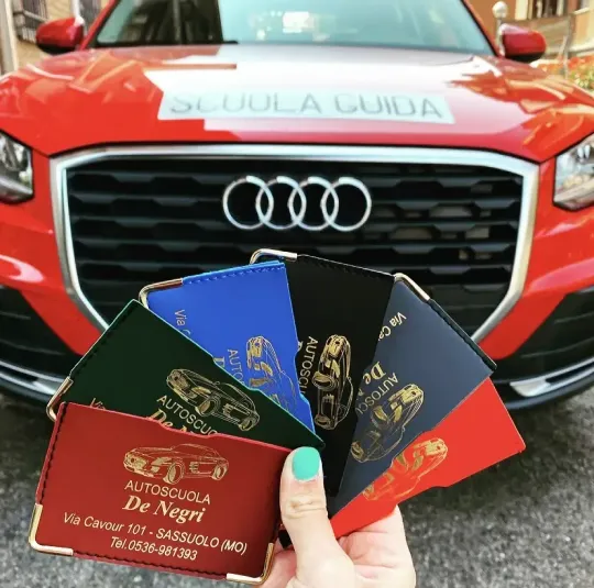 Una mano regge un mazzo di biglietti da visita colorati dell'Autoscuola De Negri davanti a un'auto della scuola guida Audi.
