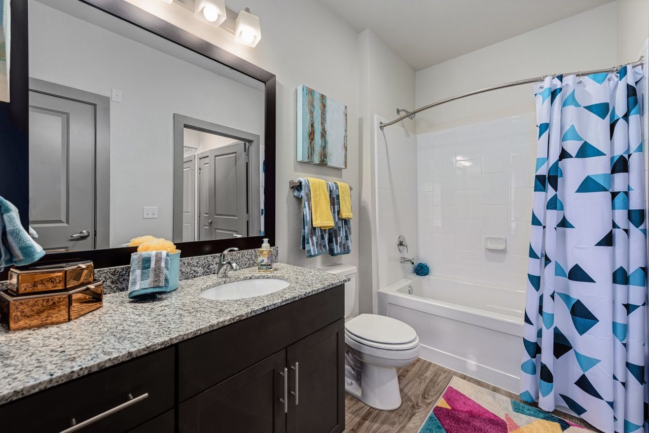 Modern apartment bathroom featuring a granite countertop vanity, sink, toilet, and a white tub with a blue geometric shower curtain.