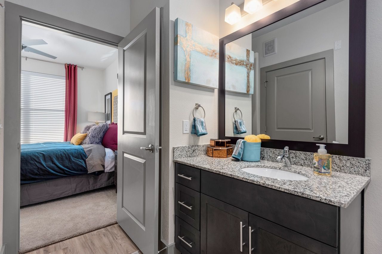 Bathroom vanity with granite countertop and large mirror; doorway leads to a bedroom with a bed.