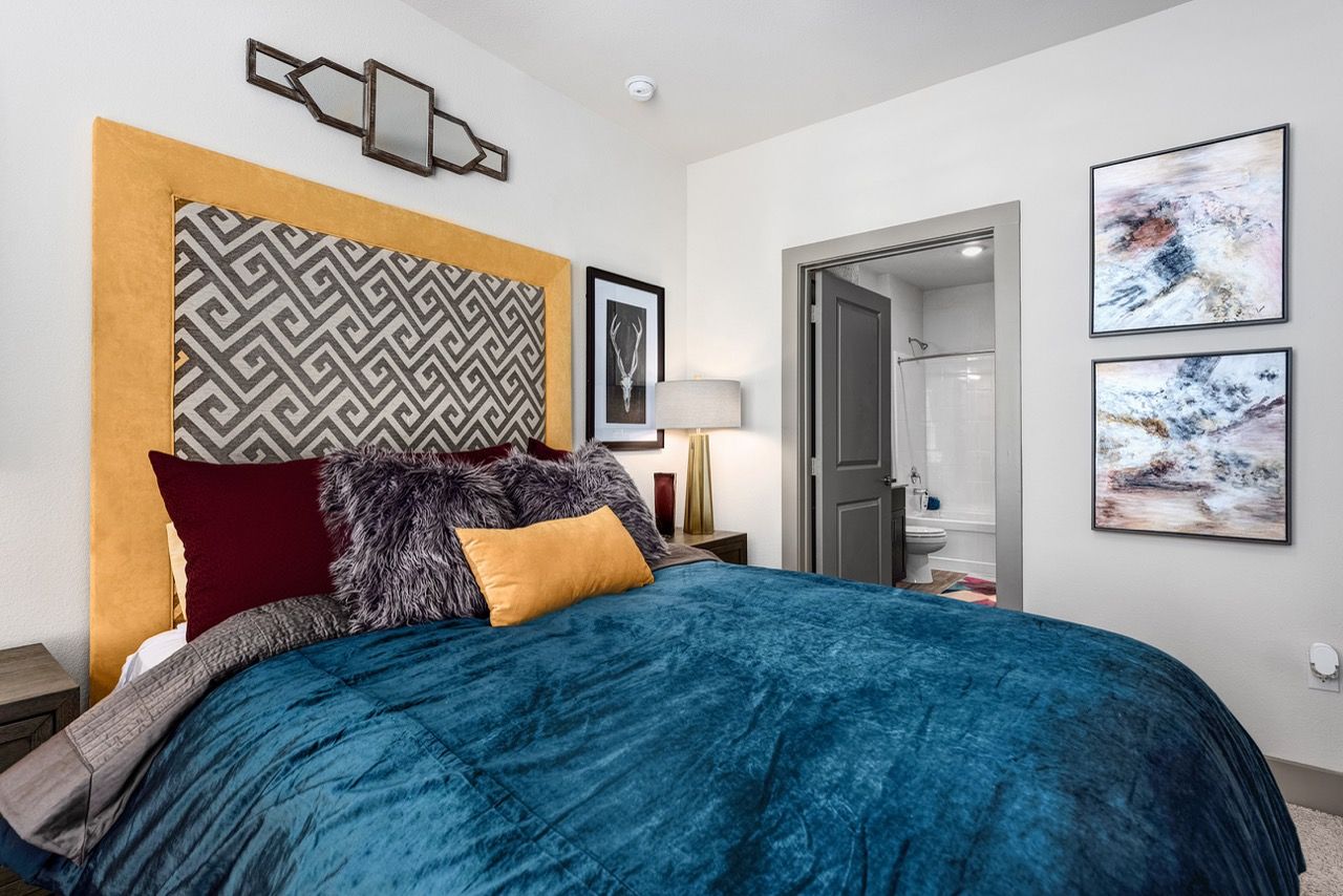 Bedroom in an apartment with a blue bedspread, bold yellow headboard, and an open doorway to the bathroom.
