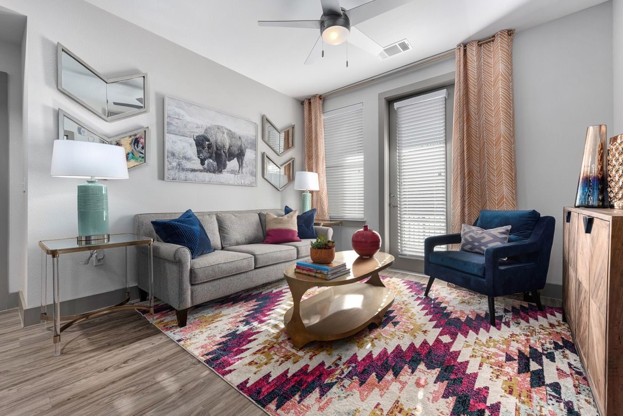 Modern living room with grey sofa, blue armchair, colorful geometric rug, and large windows.
