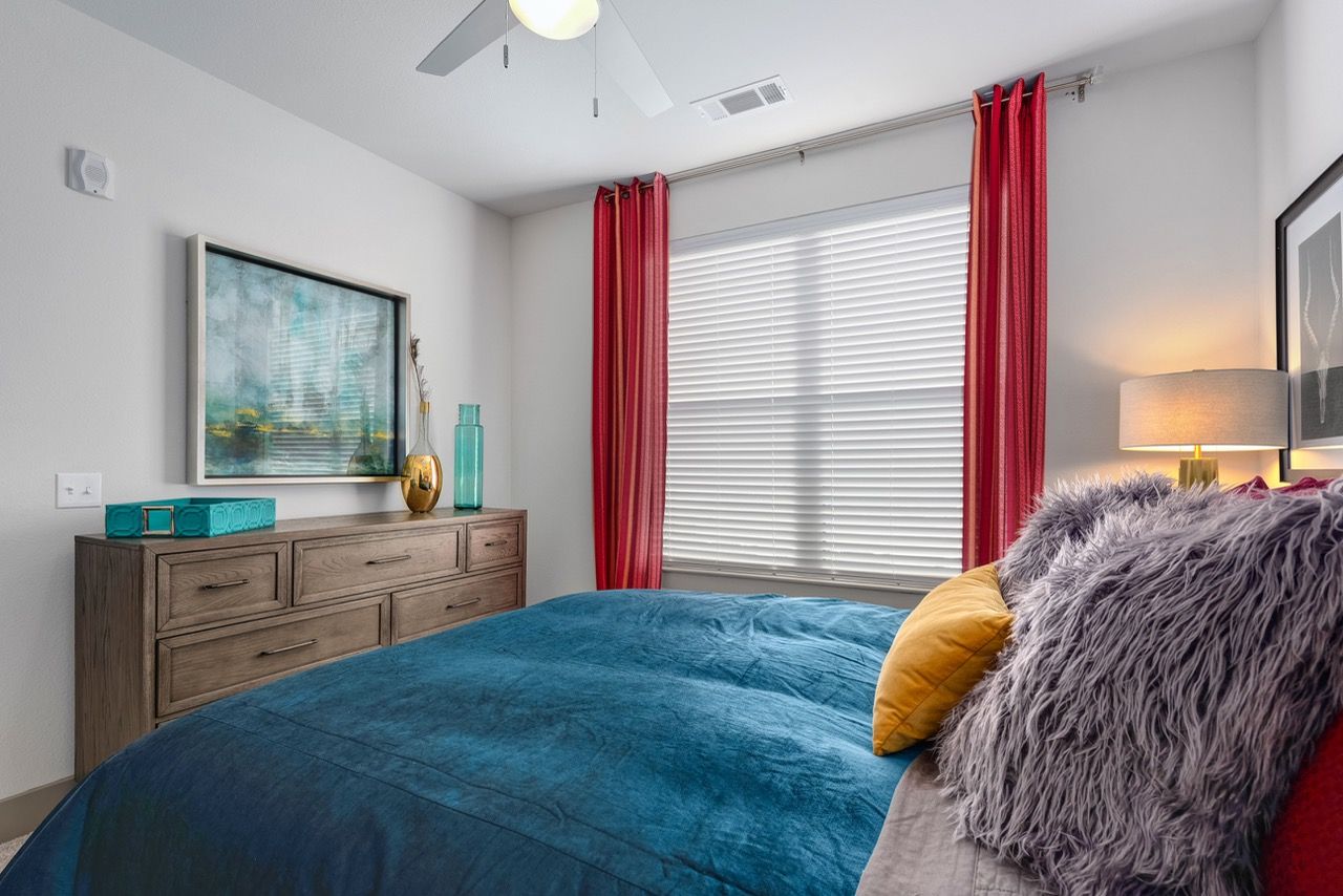 Bedroom with a blue bedspread, fluffy gray pillows, and a dresser beside a window with red curtains.