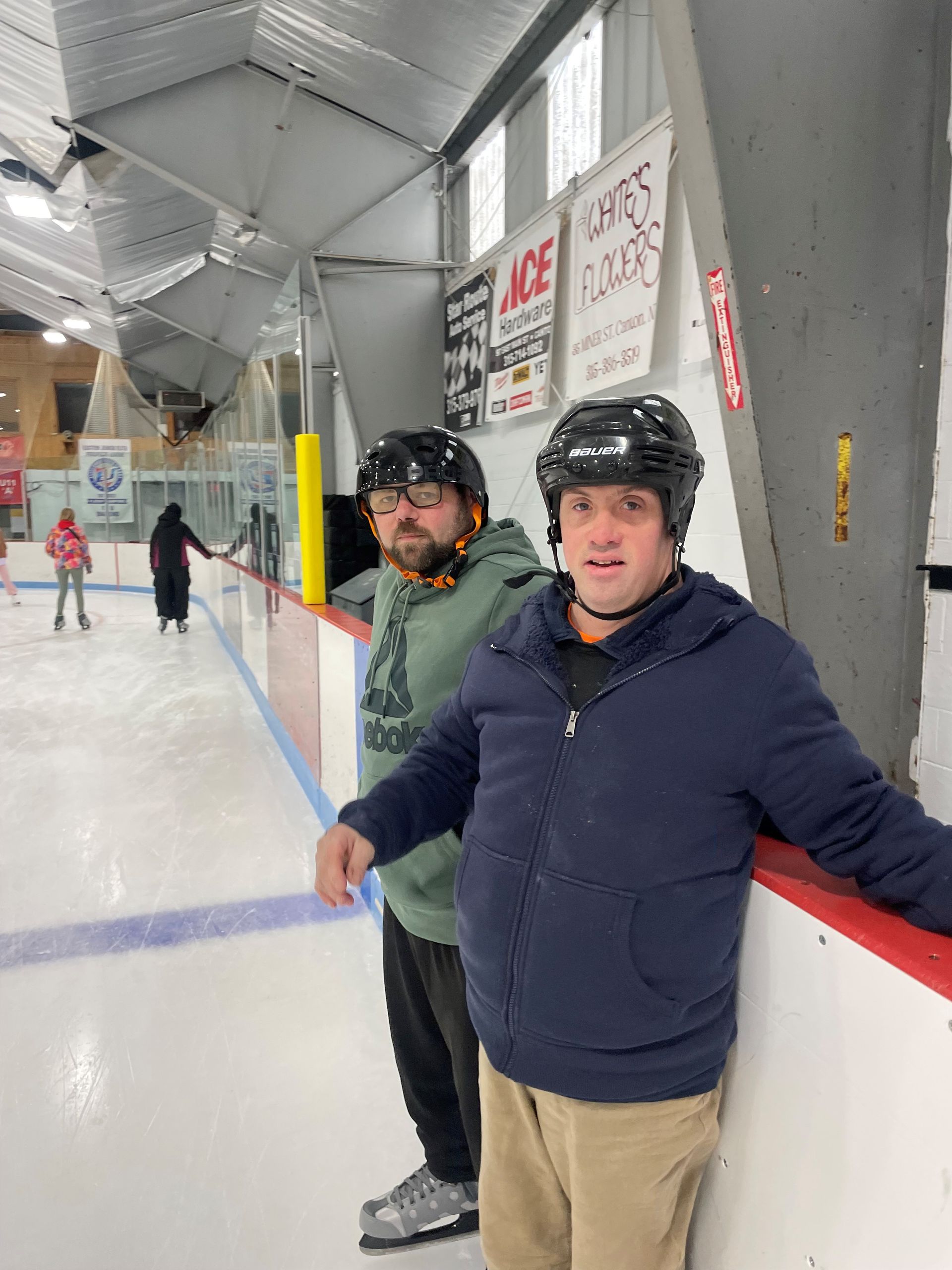 Two people standing along the boards inside an indoor ice rink, wearing helmets and casual clothing, with skaters and rink signage visible in the background.