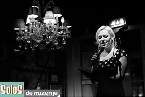 Vrouw met microfoon die voorleest uit een boek onder een kroonluchter, toneeldecor.