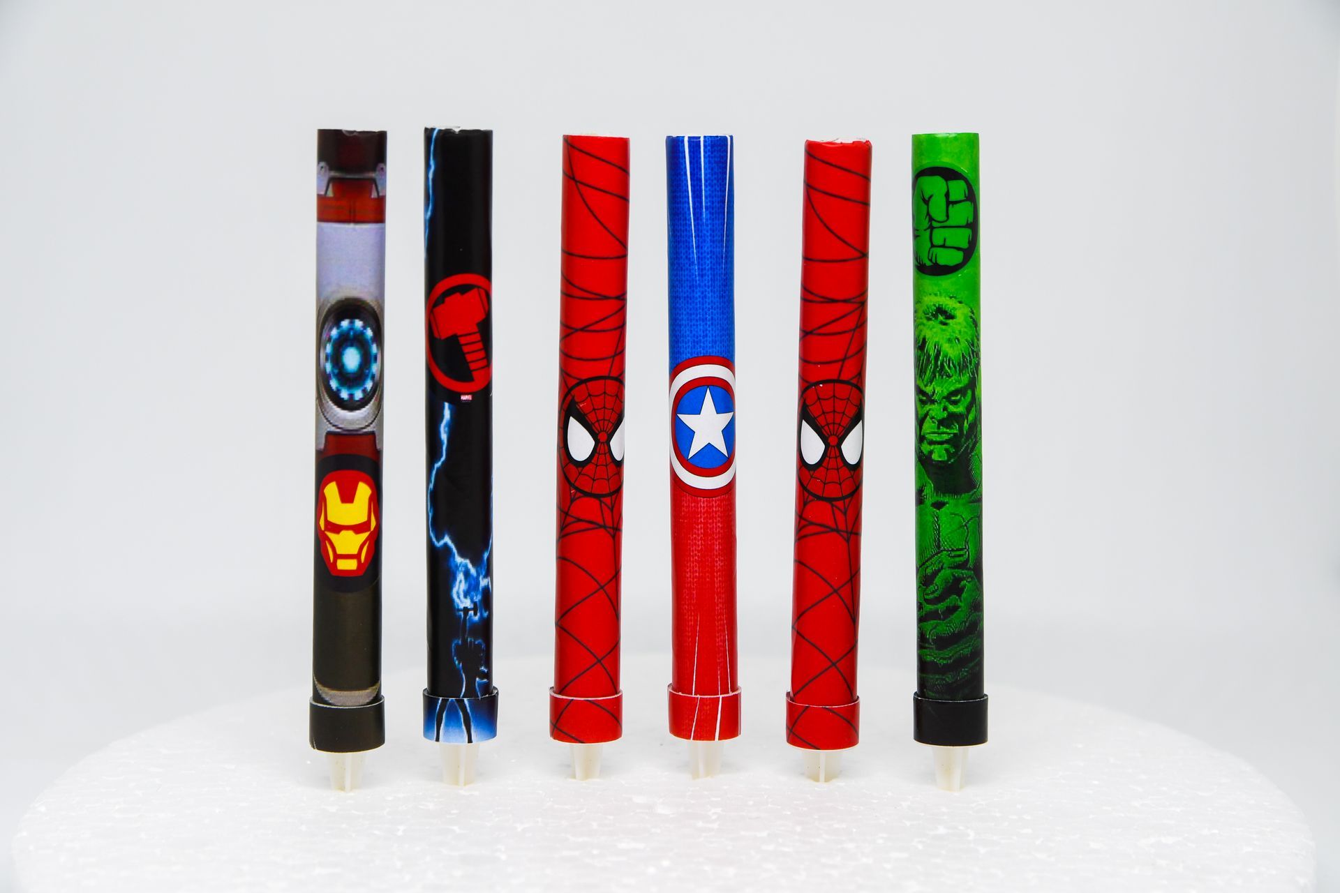 Seis velas coloridas decoradas con temática de superhéroes colocadas verticalmente sobre una superficie blanca.