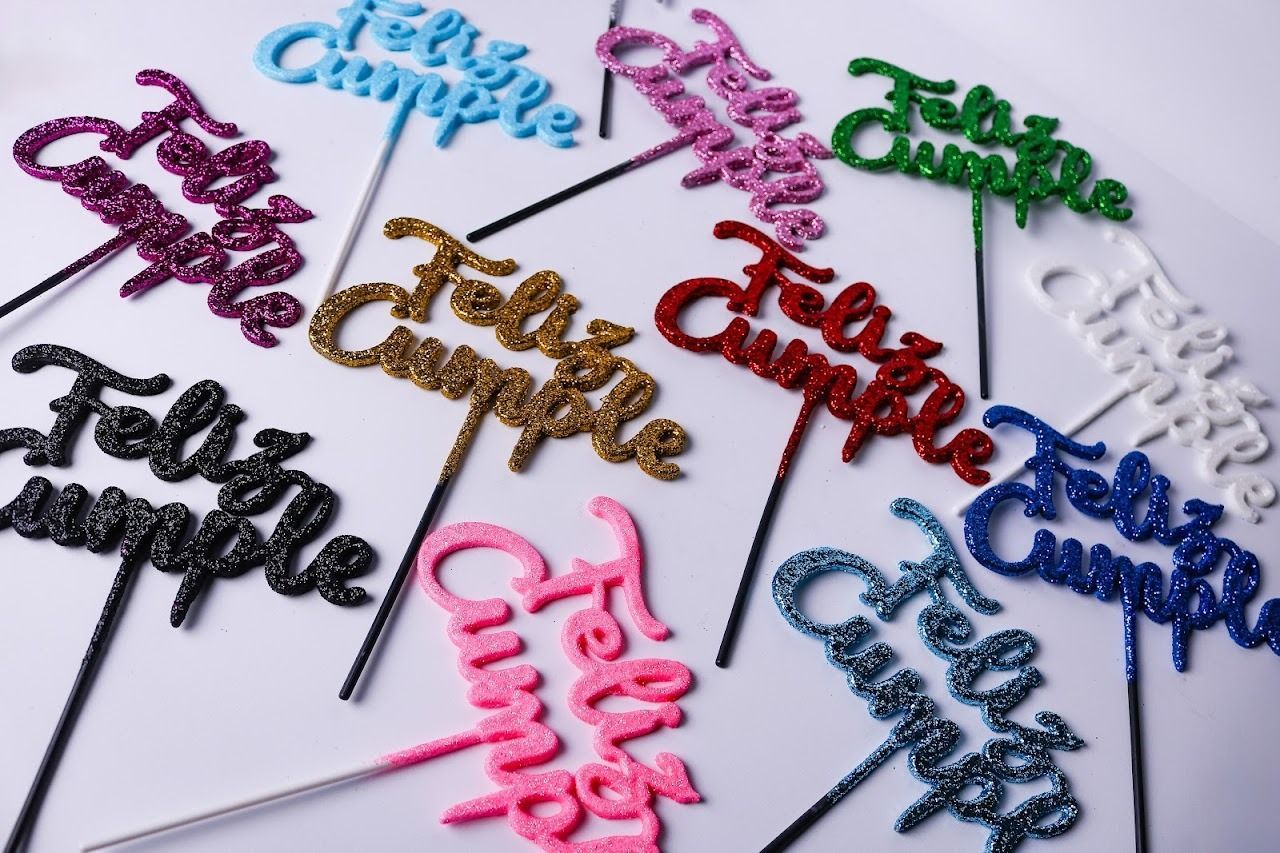 Adornos decorativos coloridos con letras para pasteles, ensartados en palitos y dispuestos sobre una superficie blanca.
