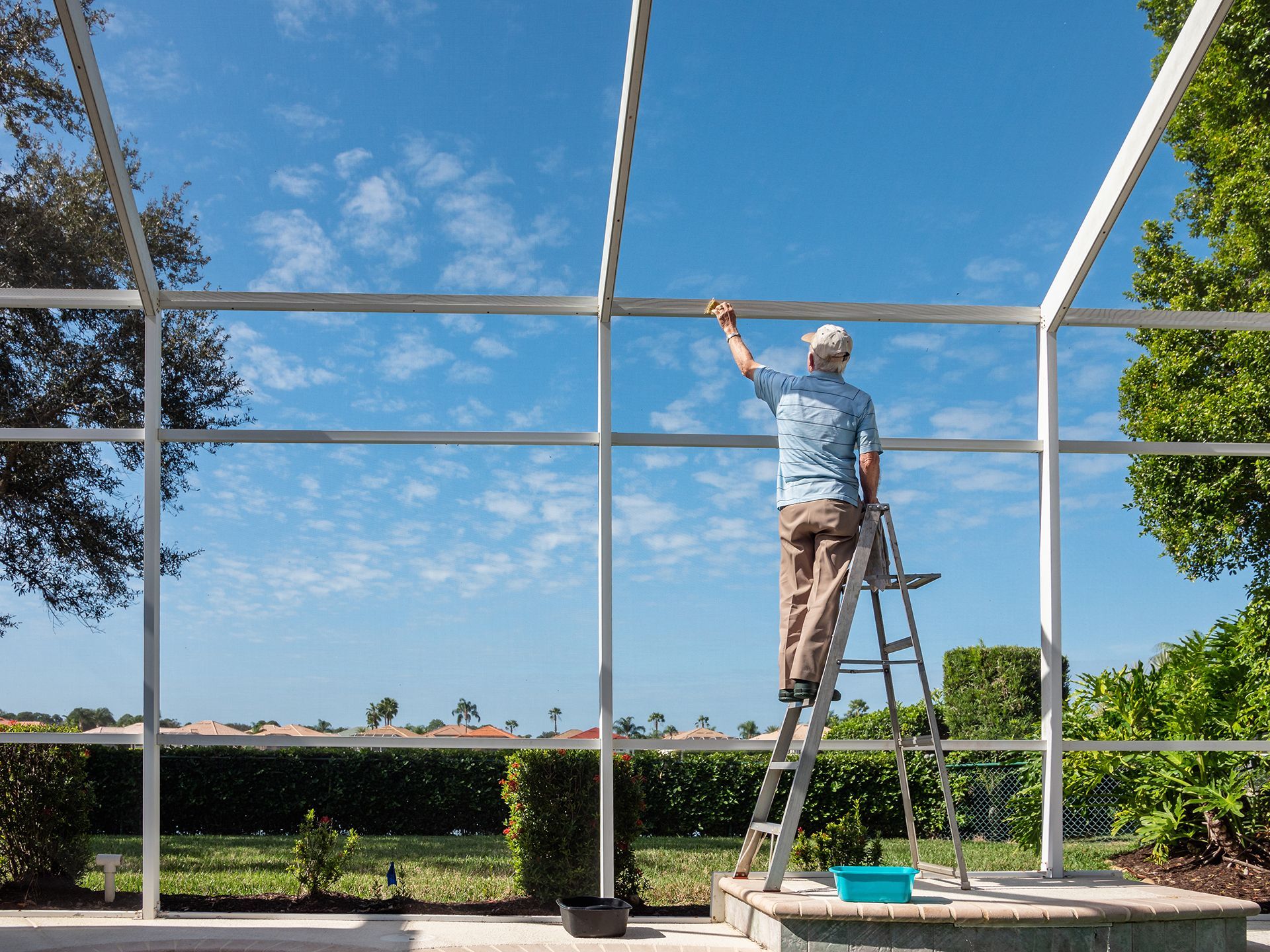 Person standing on a ladder cleaning a screened patio frame under a clear sky.