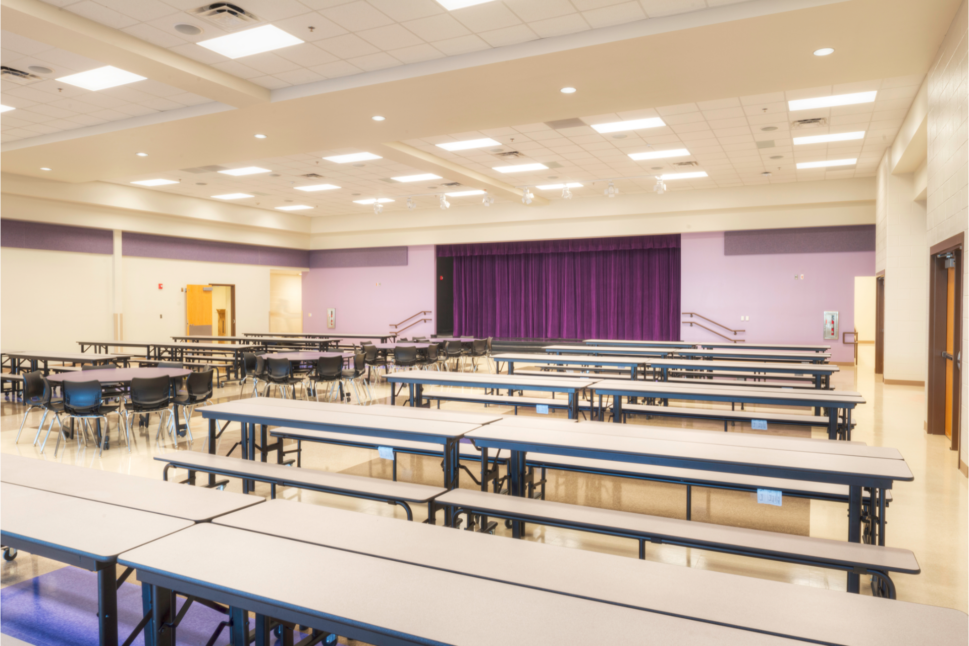 Empty cafeteria with long tables and chairs under bright ceiling lights, purple accents and curtains
