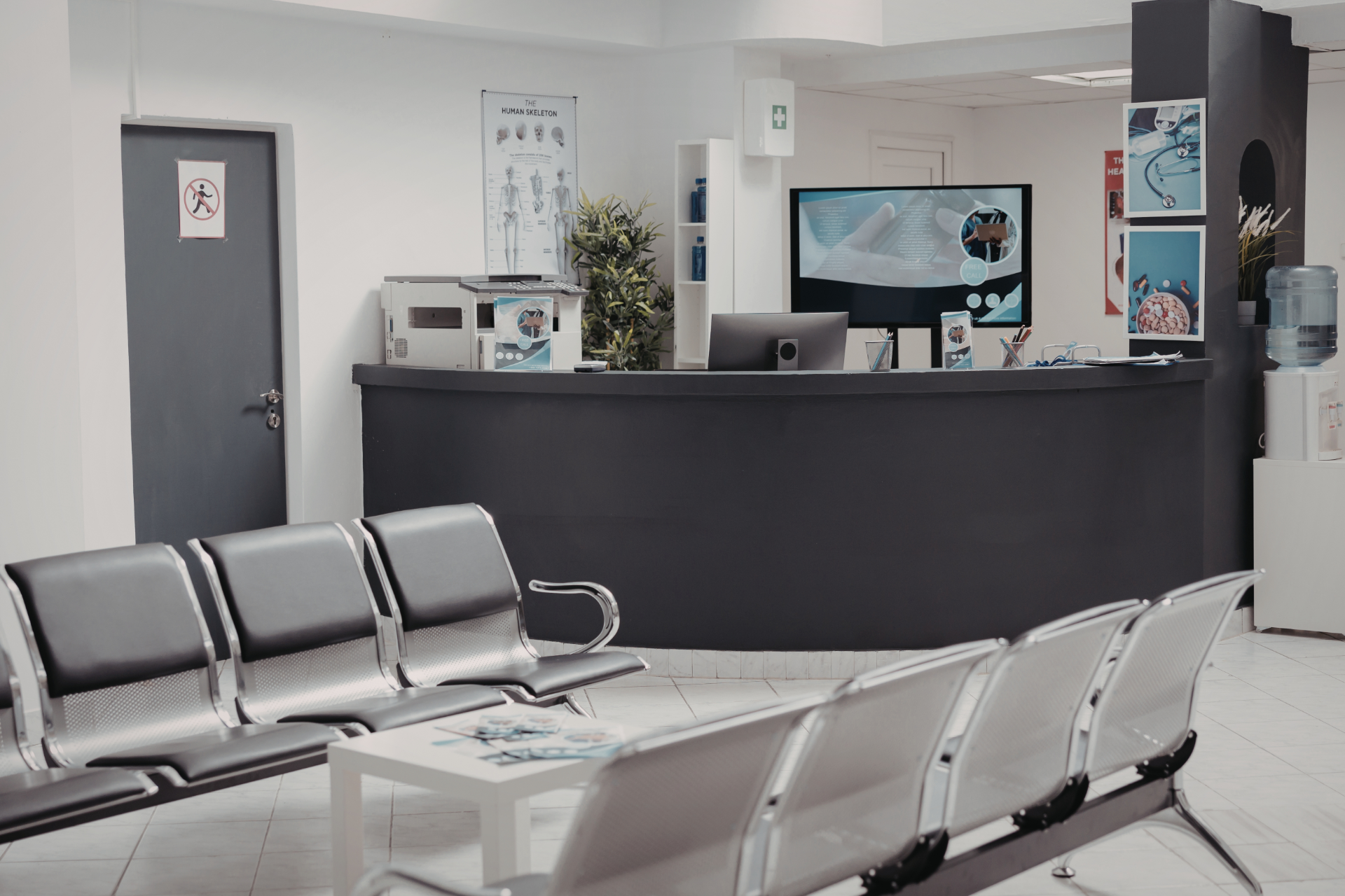 Empty medical waiting room with gray chairs, reception desk, and wall-mounted TV screens