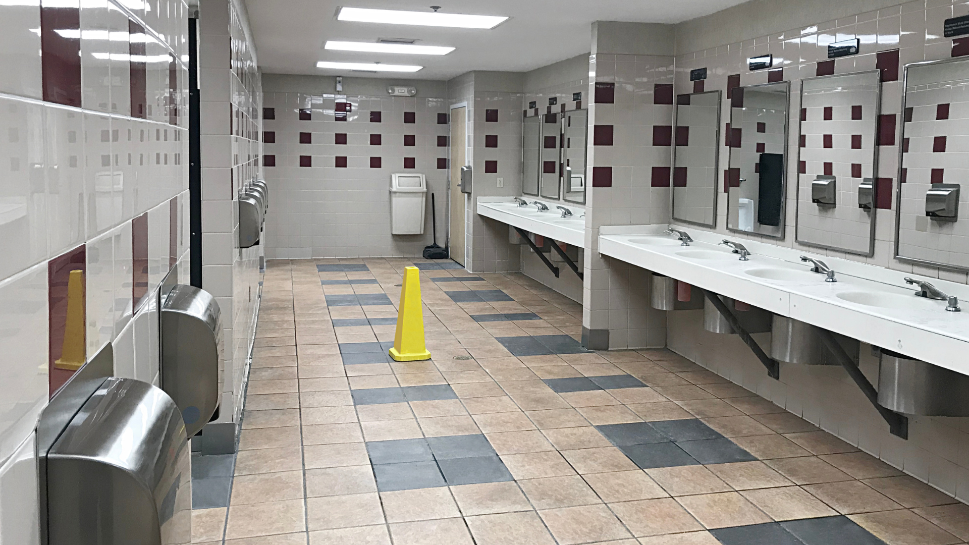 Empty public restroom with sinks, mirrors, stalls, and a yellow wet-floor sign on a tiled floor.