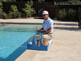 A man is kneeling next to a swimming pool with a tray of cleaning supplies